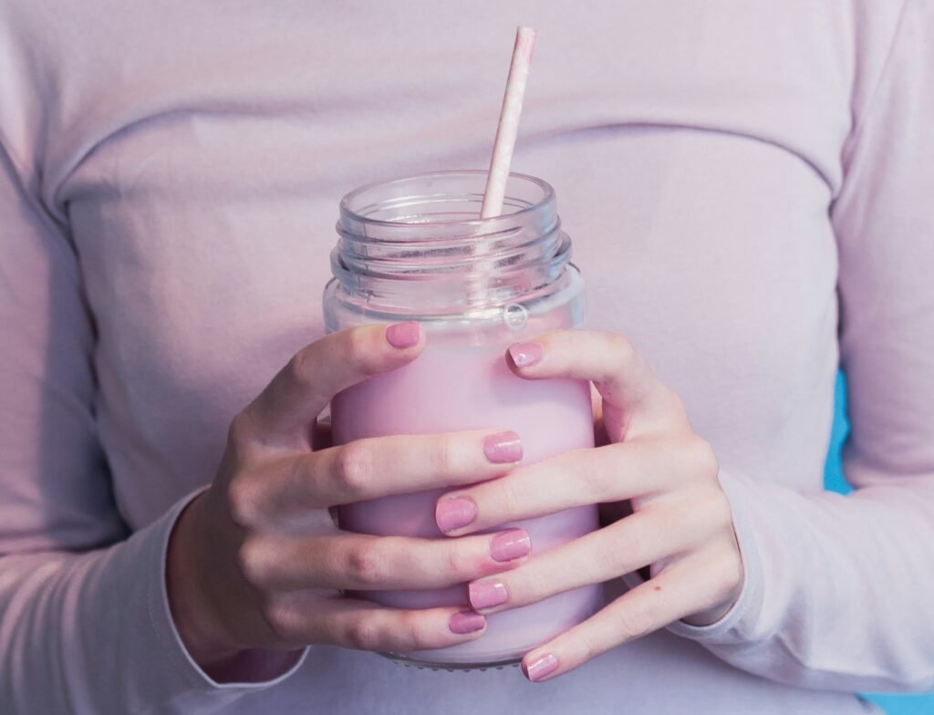 zdjęcie kobiecego torsu ubranego w różową koszulkę i rąk trzymających różowy koktajl mleczny bez laktozy w szklanym słoiku ze słomką