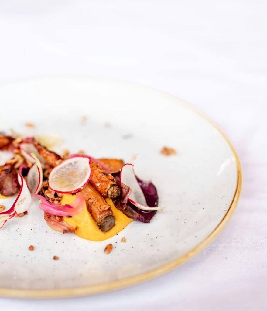 jem bez laktozy główna Elegancko podane danie bez laktozy na białym talerzu: pieczone marchewki ułożone na kremowym, żółtopomarańczowym puree. Danie udekorowane jest cienkimi plasterkami rzodkiewki, czerwoną cebulką oraz chrupiącymi elementami