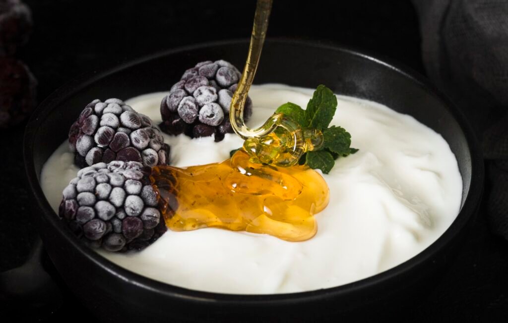 jogurt grecki Jogurt grecki bez laktozy w miseczce z dodatkiem miodu, mrożonych jeżyn i listka mięty.