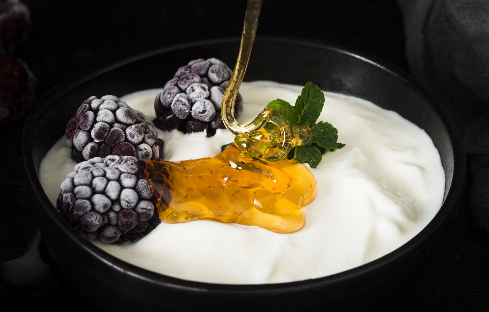 Jogurt grecki bez laktozy w miseczce z dodatkiem miodu, mrożonych jeżyn i listka mięty.