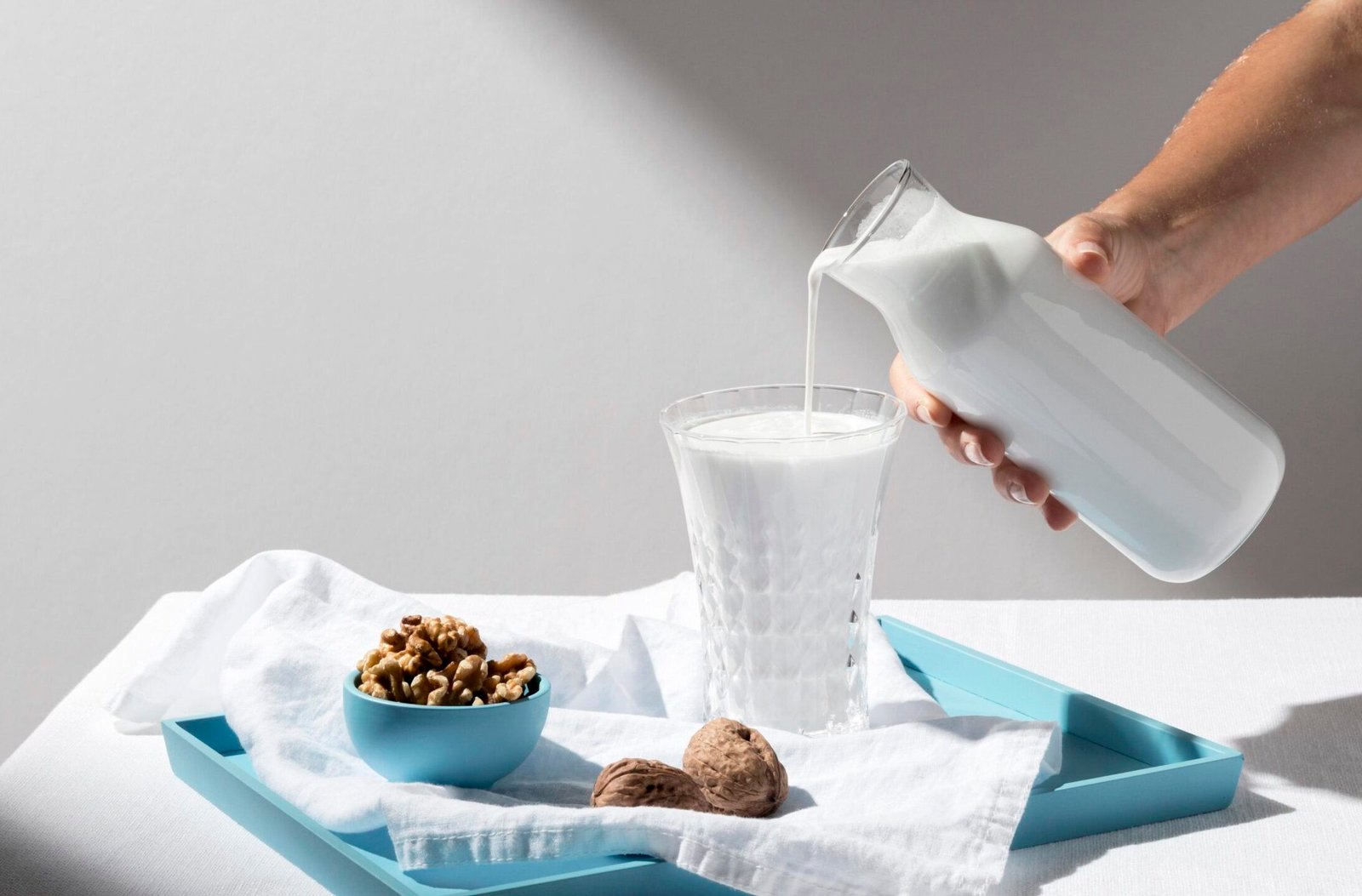 zdjęcie ręki nalewającej kefir bez laktozy z karafki do szklanki, która stoi na stole razem z miseczką orzechów włoskich i wyścieloną pod nimi białą ściereczką na niebieskiej tacy