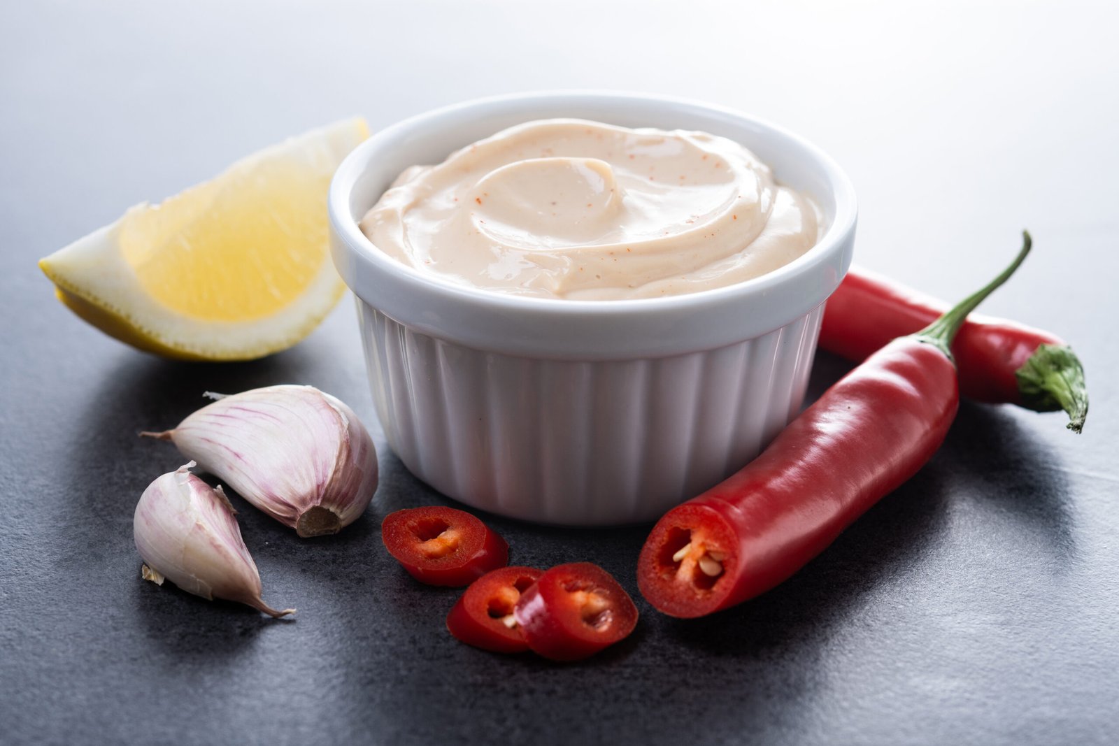 zdjęcie majonezu bez glutenu z dodatkiem przypraw w białej kokilce w otoczeniu papryczki chilli, ząbków czosnku i ćwiartki cytryny