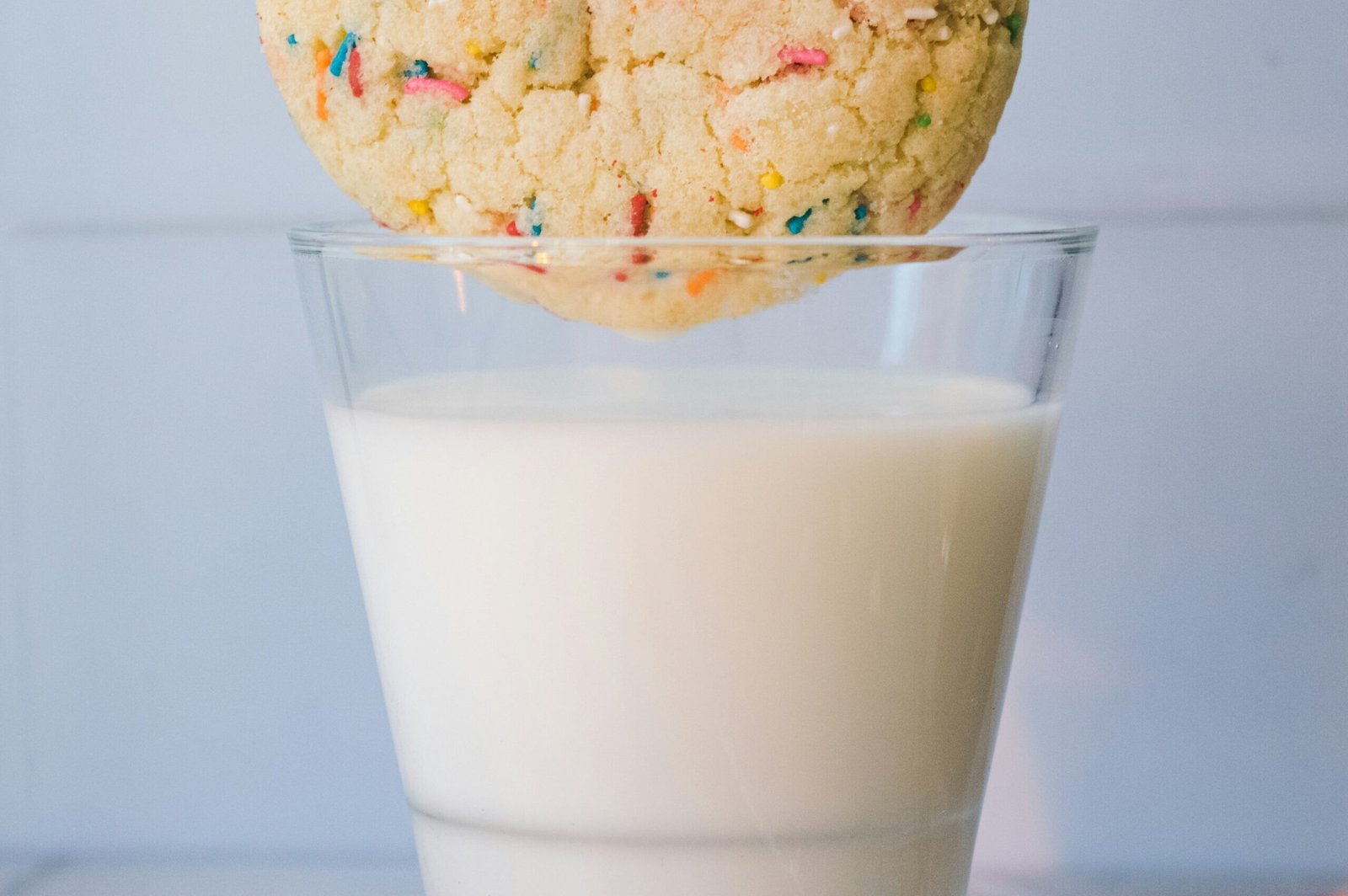 zdjęcie szklanki z mlekiem nad którą jest kruche ciasteczko z kolorową posypką, wszystko na niebieskim tle