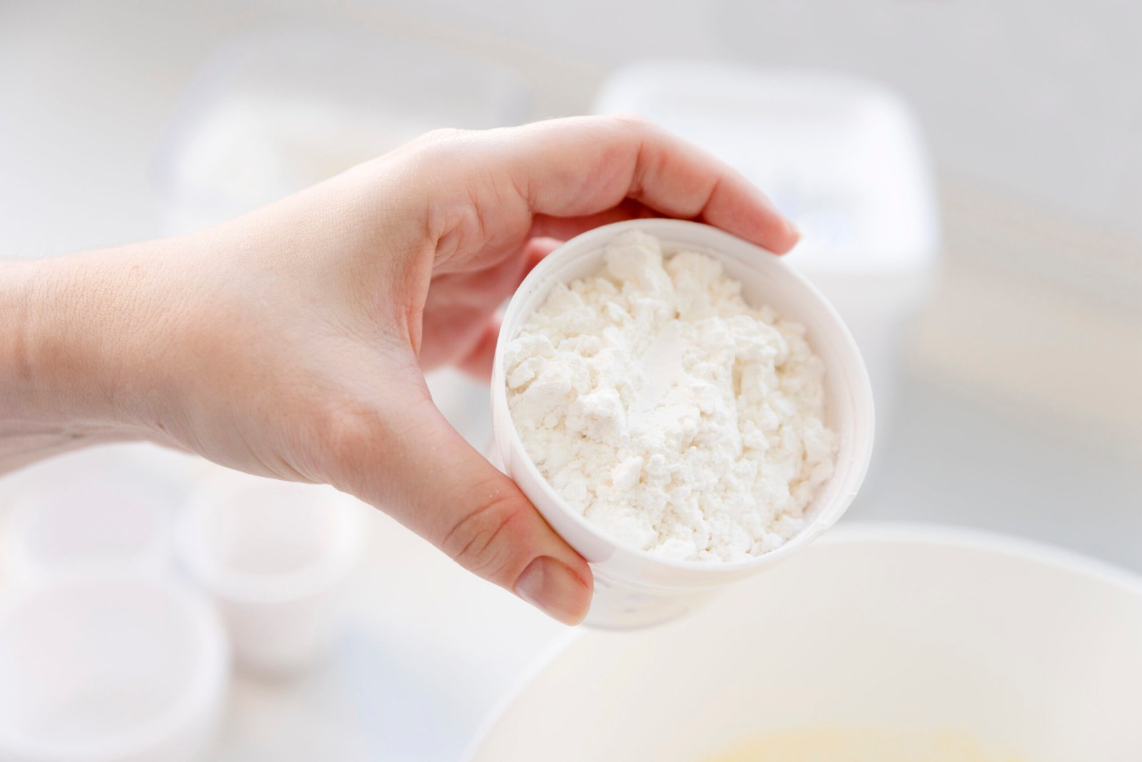 Ręka trzymająca biały kubeczek wypełniony mlekiem w proszku, na tle kuchennego blatu.