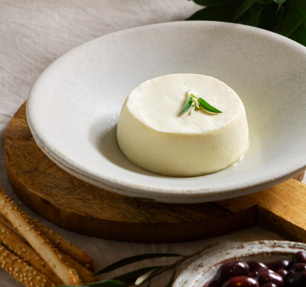 ricotta zdjęcie sera ricotta na białym talerzu udekorowanego listkiem rozmarynu, obok leżą na stole wiśnie w białej miseczce oraz paluchy pszenne