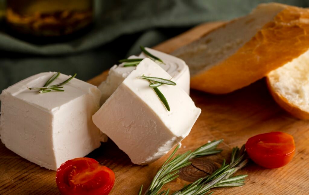 Kostki białego sera koziego z gałązkami rozmarynu, podane na drewnianej desce obok kromki chleba i pomidorków koktajlowych