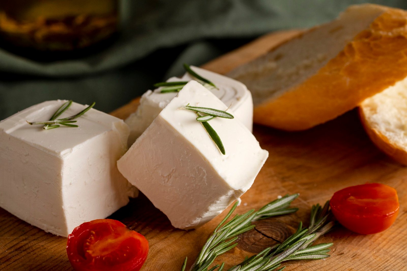 Kostki białego sera koziego z gałązkami rozmarynu, podane na drewnianej desce obok kromki chleba i pomidorków koktajlowych