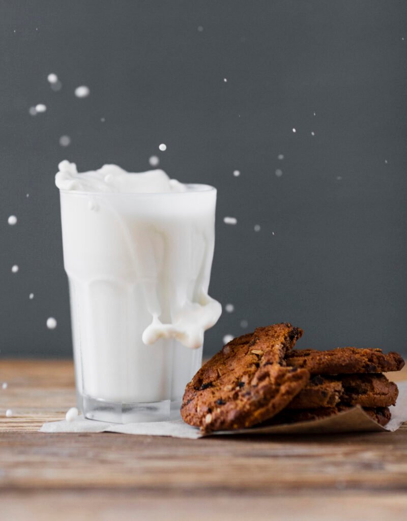 zdjęcie szklanki mleka bez laktozy wylewającego się poza jej wnętrze obok czekoladowych ciasteczek ułożonych na drewnianym stole
