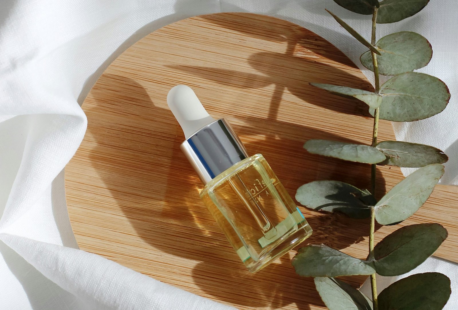 Butelka z pipetą wypełniona olejkiem kosmetycznym, leżąca na okrągłej drewnianej desce, obok gałązki eukaliptusa, w naturalnym świetle.