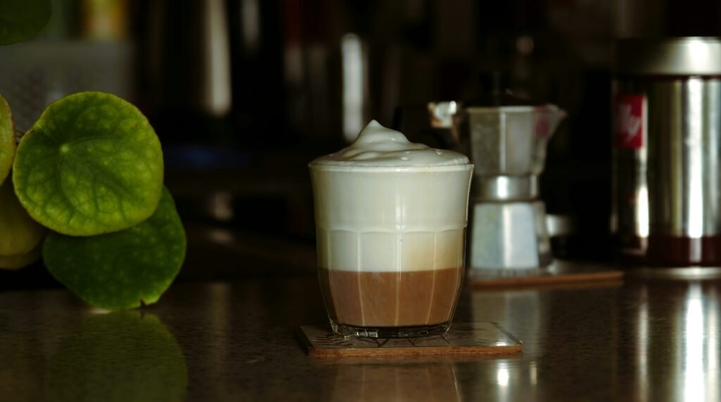 Jak zrobić mleko roślinne w domu_ Szklanka kawy latte z puszystą pianką mleczną, ustawiona na blacie kuchennym obok zielonej rośliny i kawiarki
