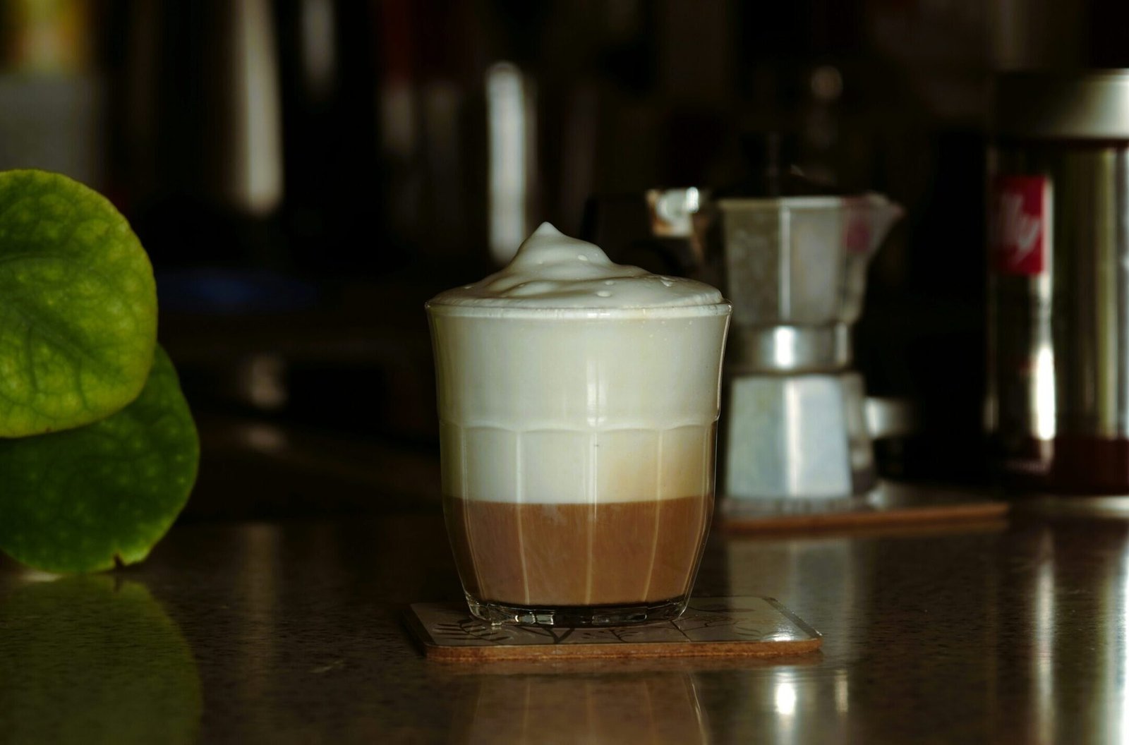 Szklanka kawy latte z puszystą pianką mleczną, ustawiona na blacie kuchennym obok zielonej rośliny i kawiarki