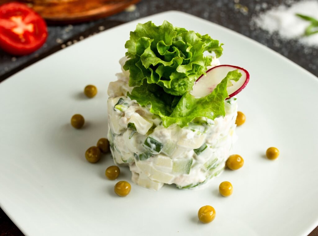 sałatka warzywna z sosem bez laktozy, z liściem sałaty i rzodkiewką na górze, podane na białym talerzu, posypanym zielonym groszkiem